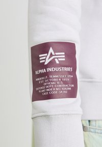 Hvit sweatshirt med en maroon etikett på ermet. Etiketten har teksten "ALPHA INDUSTRIES" og ulike kontraktdetaljer i hvitt.
