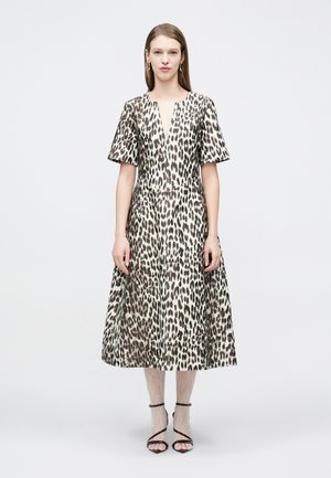 Moteris stovi vilkėdama midi suknelę su leopardų raštu ir trumpomis rankovėmis, dėvi baltas nėriniuotas pėdkelnes ir juodus basutus su dirželiais ant paprasto fono.