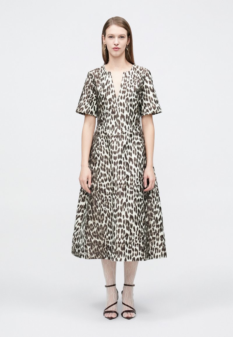 Γυναίκα που φοράει midi φόρεμα με λεοπάρ σχέδιο και κοντά μανίκια, συνδυασμένο με λευκά καλσόν από δαντέλα και μαύρες σανδάλες με λουράκια, σε απλό φόντο.