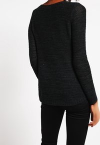 Svart stickad tröja med texturerad tygkvalitet, rund halsringning, långa ärmar och en något förlängd baksida, tillsammans med svarta byxor.