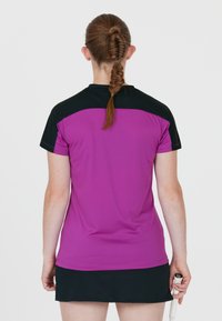 Lila idrottströja med svarta axlar, korta ärmar och en slät yta. Bärs med en svart kjol och håller en tennisracket.