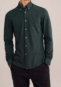 Mörkgrön skjorta med knappar framtill och långa ärmar, med en knapp-ner krage och en subtil logotyp. Tillverkad av textil med struktur och knappar fram.