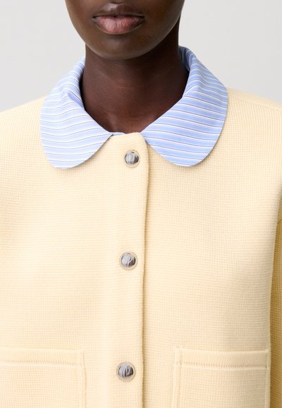 Cardigan giallo texturizzato con colletto a righe blu, bottoni grigi e due tasche frontali. Vista ravvicinata che evidenzia i dettagli.