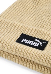 Béžová pletená čiapka s rebrovanou textúrou, ktorá má čierny obdĺžnikový štítok s nápisom "PUMA" bielymi písmenami a logom skáčucej mačky.