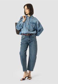 Svetlo modra denim srajca z gumbom in dvema žepoma na prsih, v kombinaciji s sproščenimi temno modrimi džinsu, ki imajo subtilne pike, in sandalčki z odprto konico.