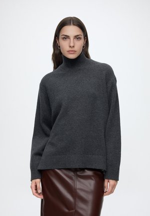 Jeune femme aux longs cheveux foncés portant un pull à col roulé gris foncé et une jupe en cuir marron sur un fond blanc uni.