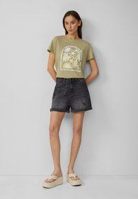 T-shirt vert olive avec un design graphique blanc, associé à un short en jean foncé avec des revers. Sandales à plateforme beige avec deux brides.