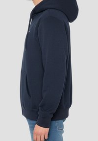 Homme portant un sweat à capuche bleu marine avec poche avant et un jean bleu clair, présenté en profil latéral sur un fond uni.