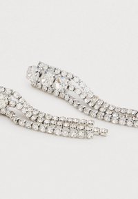 Brincos em tom prateado com três grandes pedras preciosas transparentes na parte superior e três correntes pendentes de pequenas pedras de strass transparentes.