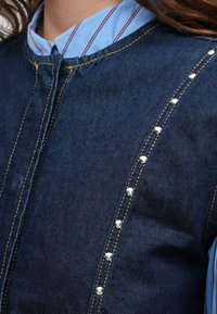 Džínsová vesta v tmavomodrej farbe so striebornými nitmi a žltým prešívaním na prednej strane. Nosená cez svetrovo svetlomodrý a červený pruhovaný tričko.