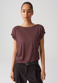 Bordeauxrotes Kurzarmoberteil mit einem geometrischen Muster in Weiß und Pink, das einen weit geschnittenen Ausschnitt und einen lockeren Sitz hat, kombiniert mit schwarzen Hosen.