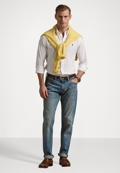 Camisa branca de botões, jeans azul claro, suéter amarelo de malha sobre os ombros, sapatos de couro castanho, relógio prateado.