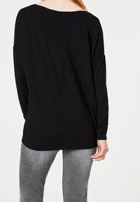 Svart långärmad topp med en bred halsringning och rak passform, med en söm längs ryggen. Matchad med grå denimbyxor.