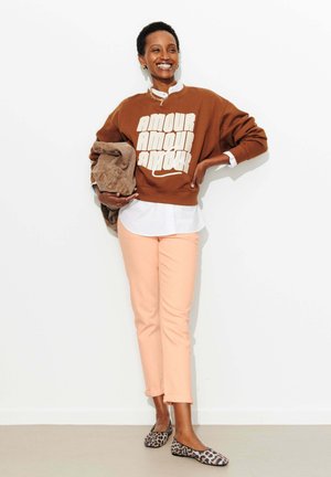 Femme souriante portant un sweat-shirt marron "AMOUR", une chemise blanche, un pantalon pêche, des ballerines léopard, tenant une veste marron pliée sur un fond blanc.