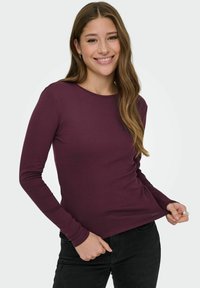 Langärmeliges, tailliertes Oberteil aus dunkelburgunderfarbenem Stoff, mit rundem Ausschnitt, glatter Textur, kombiniert mit schwarzen Hosen. Geeignet für legere Anlässe.