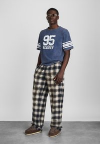 Blue cotton oversized t-shirt with white graphic and striped sleeves, paired with beige and navy checked pants and brown slip-on shoes.