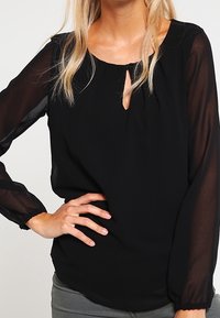 Czarna bluzka z przezroczystymi długimi rękawami, okrągłym dekoltem i detalem w kształcie klucza z przodu. Miękka, lekka tkanina; lekko luźny krój.