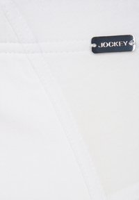 Tejido blanco con una textura suave, que presenta una etiqueta metálica plateada grabada con "JOCKEY" y un detalle de costura en contraste.