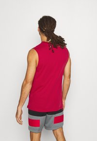 Červená bezrukávová košile v kombinaci se šedými šortkami s červenými obdélníkovými nášivkami, které prezentují neformální sportovní design. Hladký textilní materiál.