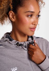 Lichtgrijze zip-up hoodie met een textuur op de oppervlakte, capuchon met trekkoorden en zwarte accenten op de schouders. Witte logo op de borst.