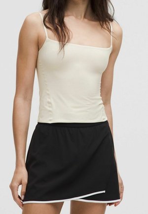 Frau trägt cremefarbenes Spaghetti-Trägertop und schwarzen Rock mit weißem Besatz, gezeigt vom Hals bis zur Mitte des Oberschenkels vor grauem Hintergrund.