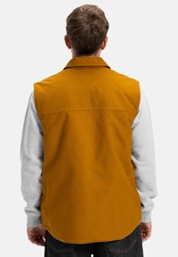 Gilet senza maniche di colore giallo senape, con colletto e una texture liscia, abbinato a una camicia grigia a maniche lunghe sotto.