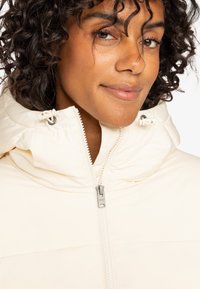 Veste en duvet crème avec un col montant, fermeture éclair visible et ajustements par cordons. Texture douce, avec un design ajusté et près du corps.