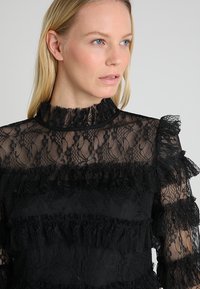 Svart blonde topp med høy hals, volanger og lagdelte teksturer. Stoffet er gjennomskinnelig med intrikate blomsterdesign.