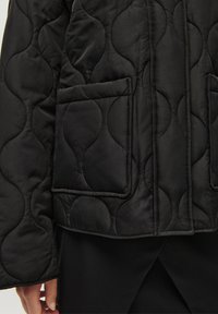 Black quilted jacket featuring a wavy texture, two front pockets, and a straight cut. The fabric appears soft and slightly glossy.