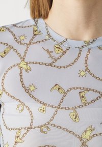 Kirkas sininen paita, jossa on toistuva keltainen ketjumalli, johon kuuluu saappaita, sydämiä, hevosenkenkiä ja tähtiä. Pyöreä pääntie, kevyt kangas.