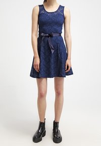 Robe en dentelle bleu marine avec un corsage ajusté, une jupe évasée et un nœud en ruban à la taille. Associée à des bottines noires à semelles épaisses.