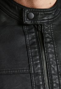 Veste en cuir noir avec un col haut, texture subtile, fermeture éclair centrale et un bouton-pression. Coutures visibles le long des bords.
