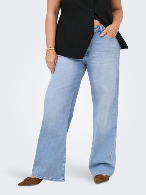 Kvinne iført lyseblå vide jeans, brune spisse sko, svart ermeløs blazer og et gullarmbånd, står med én hånd i lommen.