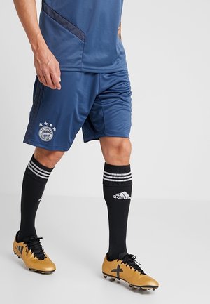 Personne portant un short de football bleu du FC Bayern Munich, des chaussettes noires Adidas avec des bandes blanches, et des chaussures de football Adidas dorées sur un fond uni.