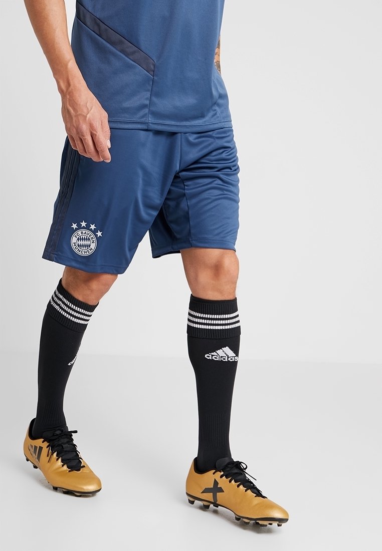 Personne portant un short de football bleu du FC Bayern Munich, des chaussettes noires Adidas avec des bandes blanches, et des chaussures de football Adidas dorées sur un fond uni.