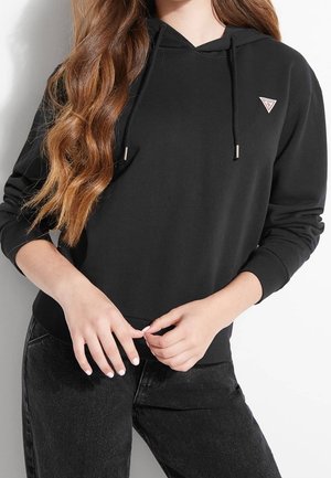 Vrouw met lang golvend haar die een zwarte hoodie draagt met trekkoorden en een klein driehoekig logo, gecombineerd met een zwarte broek, handen die de zoom aanpassen.