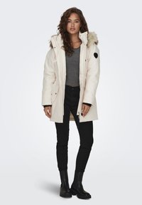 Parka jas in lichtbeige met een met bont gevoerde capuchon, sluiting met rits aan de voorkant en zijzakken, gecombineerd met zwarte skinny jeans en enkellaarsjes.