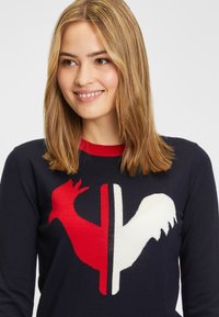 Uśmiechająca się kobieta z blond włosami, nosząca granatowy sweter z czerwono-białym wzorem podzielonego koguta z przodu.