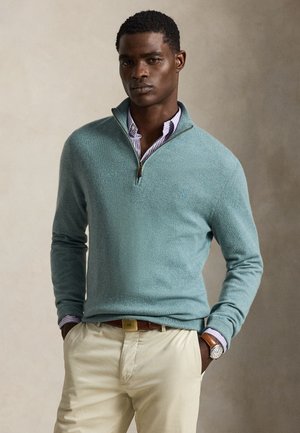 Homme portant un pull bleu clair à moitié zip par-dessus une chemise rayée, pantalon beige et une montre-bracelet, mains dans les poches sur un fond neutre.