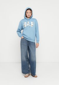 Blue hooded sweatshirt with floral logo, front pocket; paired with wide-leg faded denim jeans and patterned slip-on shoes. Plain background.