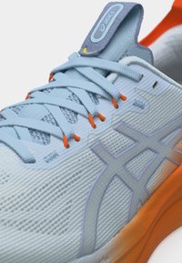 Nærbilde av lyseblå Asics-sneakers med oransje lisseringer, nettstoff på overdelen og oransje detaljer på sålen.