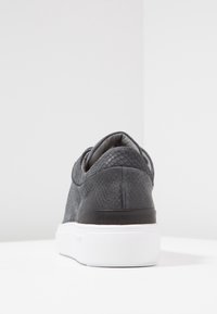 Svart texturerade sneakers med ett orm-skinne-mönster, vit gummisul, och vadderad krage, som visar upp mjuka sömmar och en rundad bakdel.