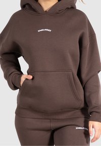 Felpa con cappuccio marrone realizzata in morbido tessuto, con tasca frontale e logo bianco "SMILODOX". Vestibilità rilassata con polsini a costine.