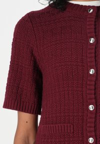 Cardigan a maglia bordeaux con maniche corte, tessuto strutturato, collo e orlo a coste, bottoni argentati e dettaglio con tasca frontale.