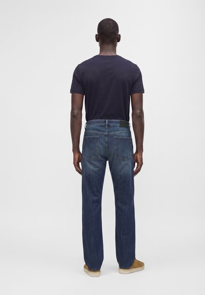 Homme debout de dos portant un jean bleu foncé, un t-shirt noir et des chaussures marron clair sur un fond gris uni.
