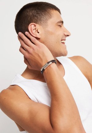 Pulsera de cuero negro con detalles metálicos plateados, llevada en la muñeca. El modelo muestra un primer plano del brazo y la mano tocando el cuello.