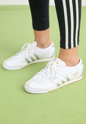 Nohy v bielych teniskách Adidas Italia so striebornými pruhmi a šnúrkami stoja na svetlozelenej textúrovanej ploche.