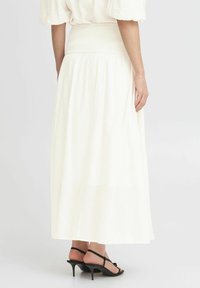 Jupe longue blanche fluide avec une taille smockée, faite d'un tissu léger, associée à des sandales à talons noires. Texture lisse.