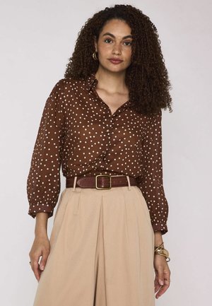 Femme aux cheveux bouclés porte un chemisier marron à pois, un pantalon beige taille haute et une ceinture marron, debout devant un fond uni.