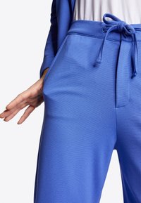 Nahaufnahme einer Person in blauen Jogginghosen mit Kordelzug, die Hand nahe der Seitentasche, ein weißes Shirt und der Ärmel einer blauen Jacke sind sichtbar.
